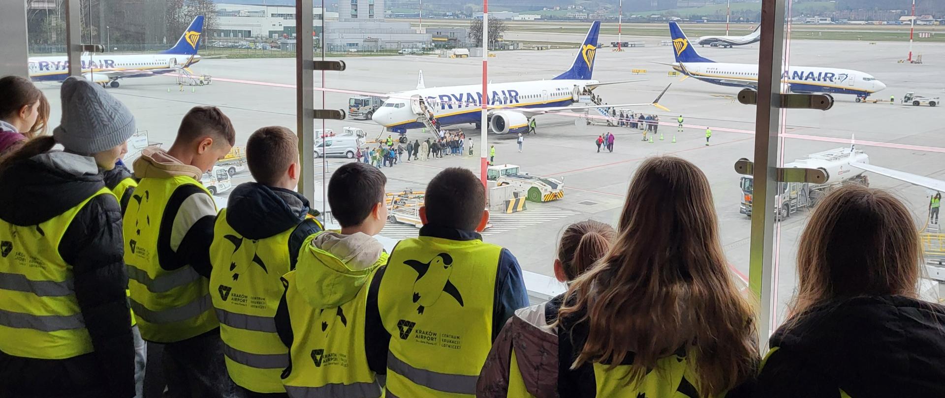 Uczniowie w żółtych kamizelkach patrzą na płytę lotniska na której stoją samoloty