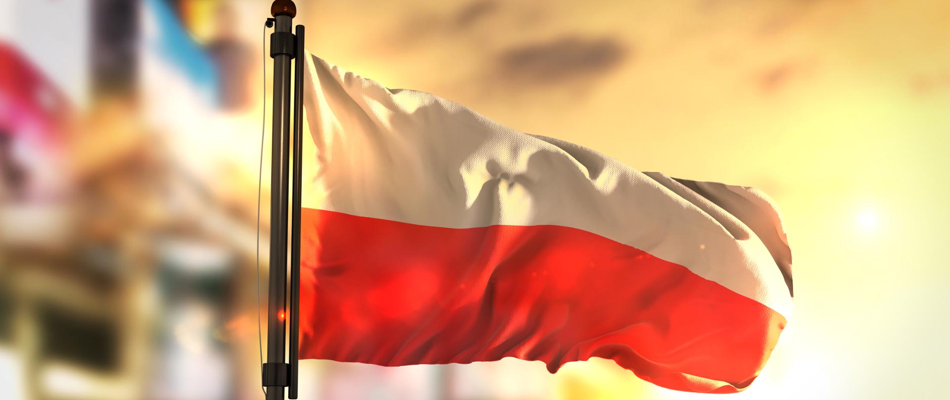Powiewająca na maszcie flaga Polski na tle zachodzącego w mieście miasta