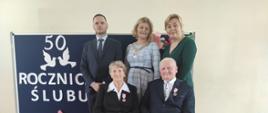 Państwo Halina i Sylwester na tle granatowej tablicy z napisem wraz z Wójtem, Z-cą Wójta i Z-cą Kierownika USC