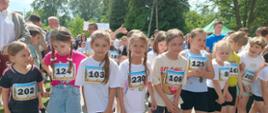 Dziewczynki w sportowych strojach z przypiętymi numerami startowymi stoją rzędem na linii startu na asfaltowej ścieżce, przygotowując się do rozpoczęcia biegu. W tle widać drzewa oraz innych uczestników wydarzenia.
