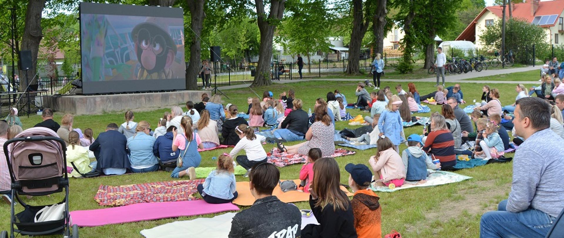 dzieci i dorośli siedzą na kocach na trawie i oglądają bajkę na dużym ekranie 