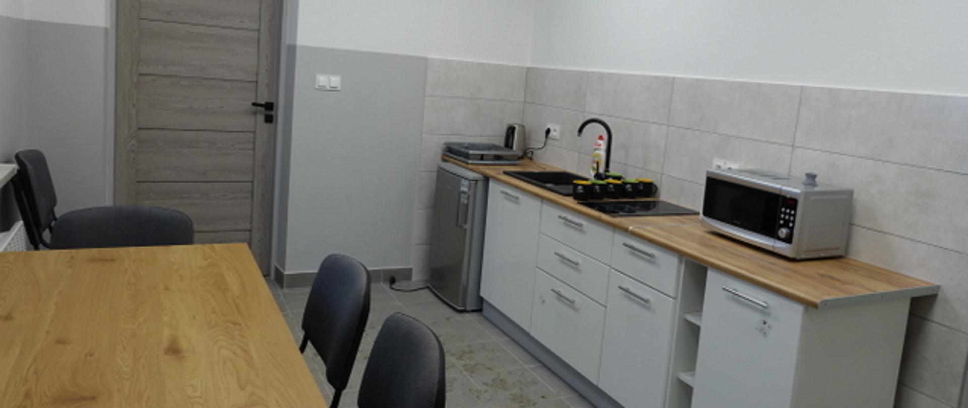 wyremontowane pomieszczenie widoczny aneks kuchenny stół lodówka krzesła kolorystyka odcieni szarości