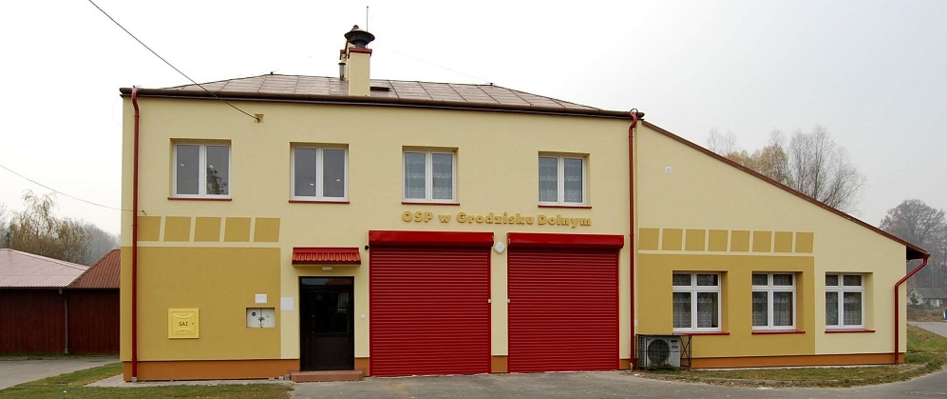 Żółty, dwupiętrowy budynek Straży Pożarnej, dwoje czerwonych dużych drzwi i jedne mniejsze brązowe. 