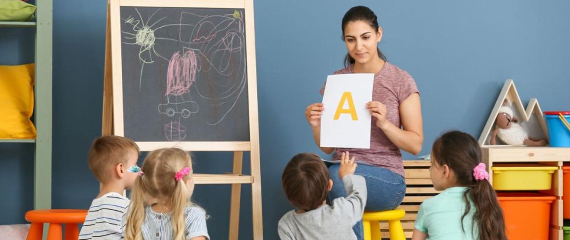 Zdjęcie sali lekcyjnej, siedząca kobieta - nauczycielka trzymająca w ręku kartkę na której widnieje litera A, przed kobietą siedzi czworo dzieci patrzących w stronę kobiety.