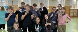 Jedenaścioro uczniów naszej szkoły, którzy brali udział w zawodach pozuje do wspólnego zdjęcia na sali gimnastycznej. Wszyscy ubrani są w stroje sportowe. Na szyjach mają medale.