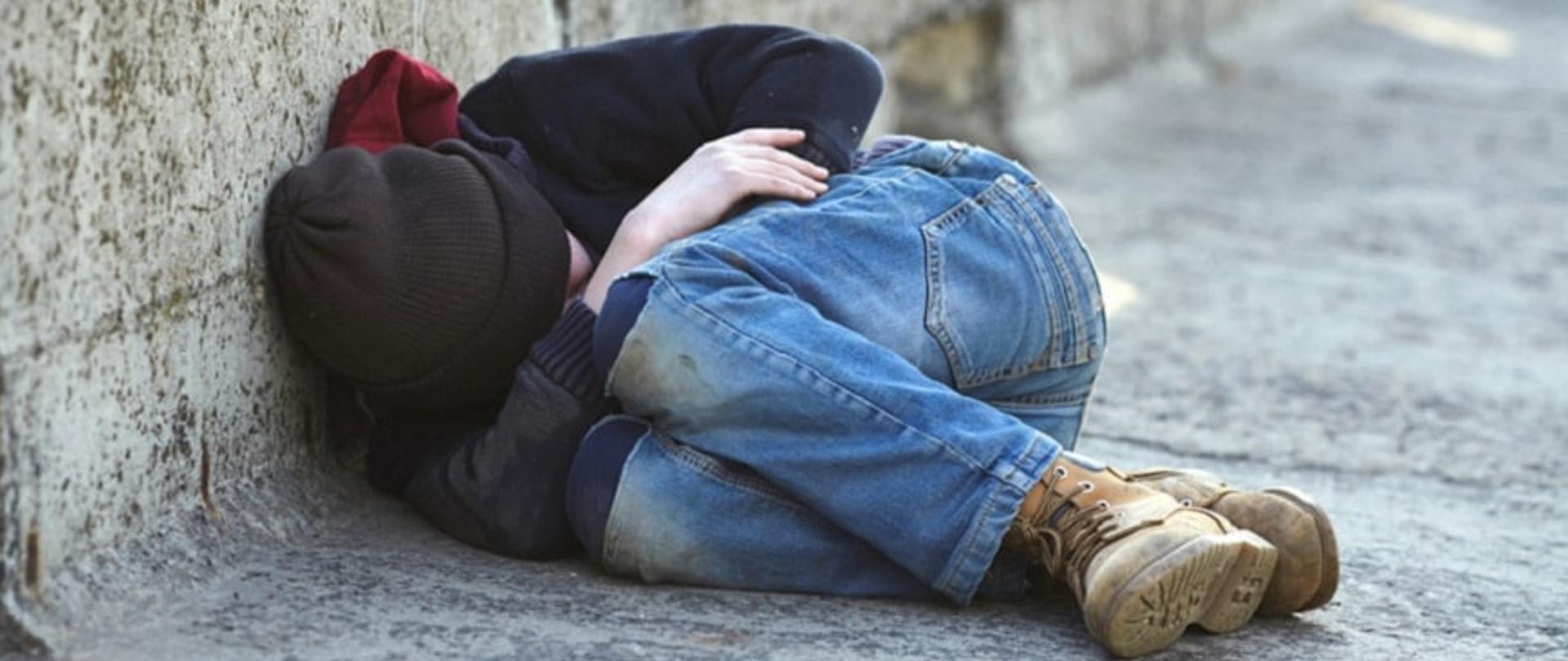 bezdomna skulona osoba leżąca pod ścianą na zewnątrz