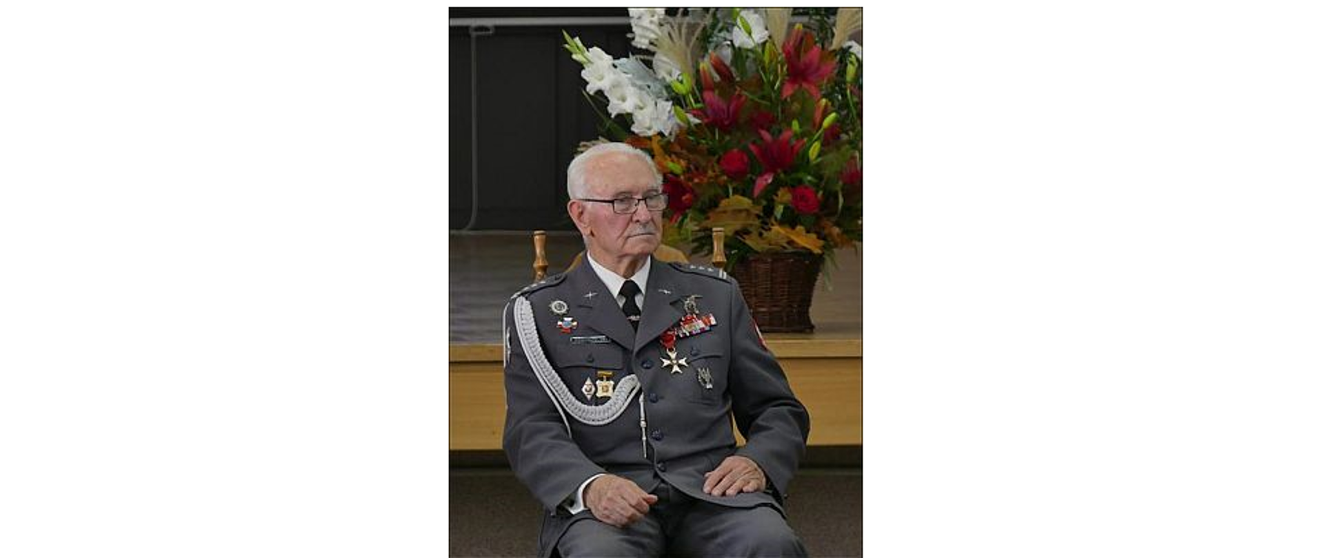 Na zdjęciu znajduje się Pułkownik pil. Kazimierz Pogorzelski ubrany w mundur z licznymi odznaczeniami. W tle scena z koszem pełnym kwiatów.