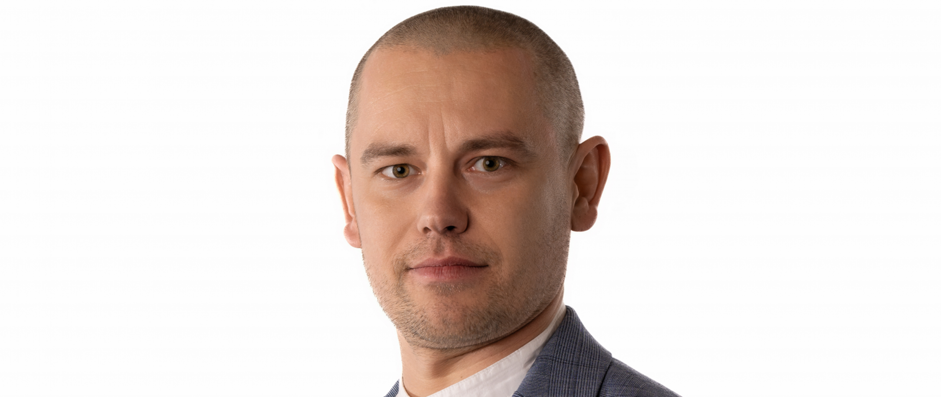 Na zdjęciu widać mężczyznę z bardzo krótko ściętymi, ciemnymi włosami. Brązowe oczy lekko zmarszczone brwi. Ubrany w szarą marynarkę i białą koszule ze stójką 