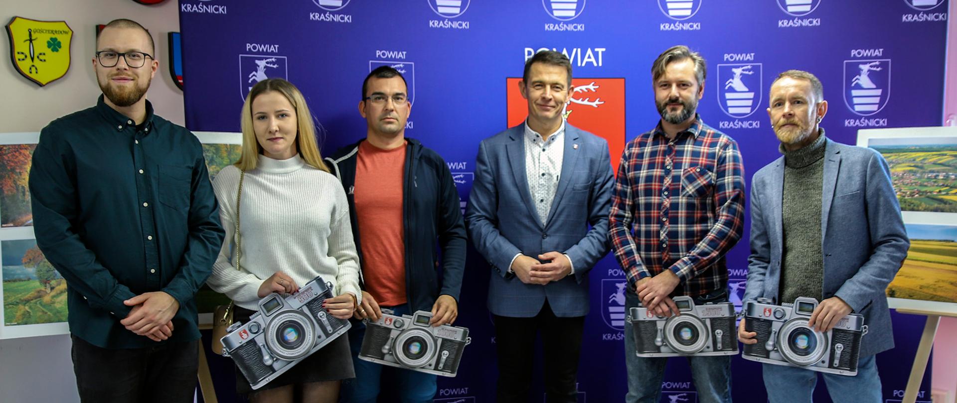 Zdjęcie grupowe laureatów konkursu. Sześć osób na tle niebieskiej ścianki Powiatu Kraśnickiego.