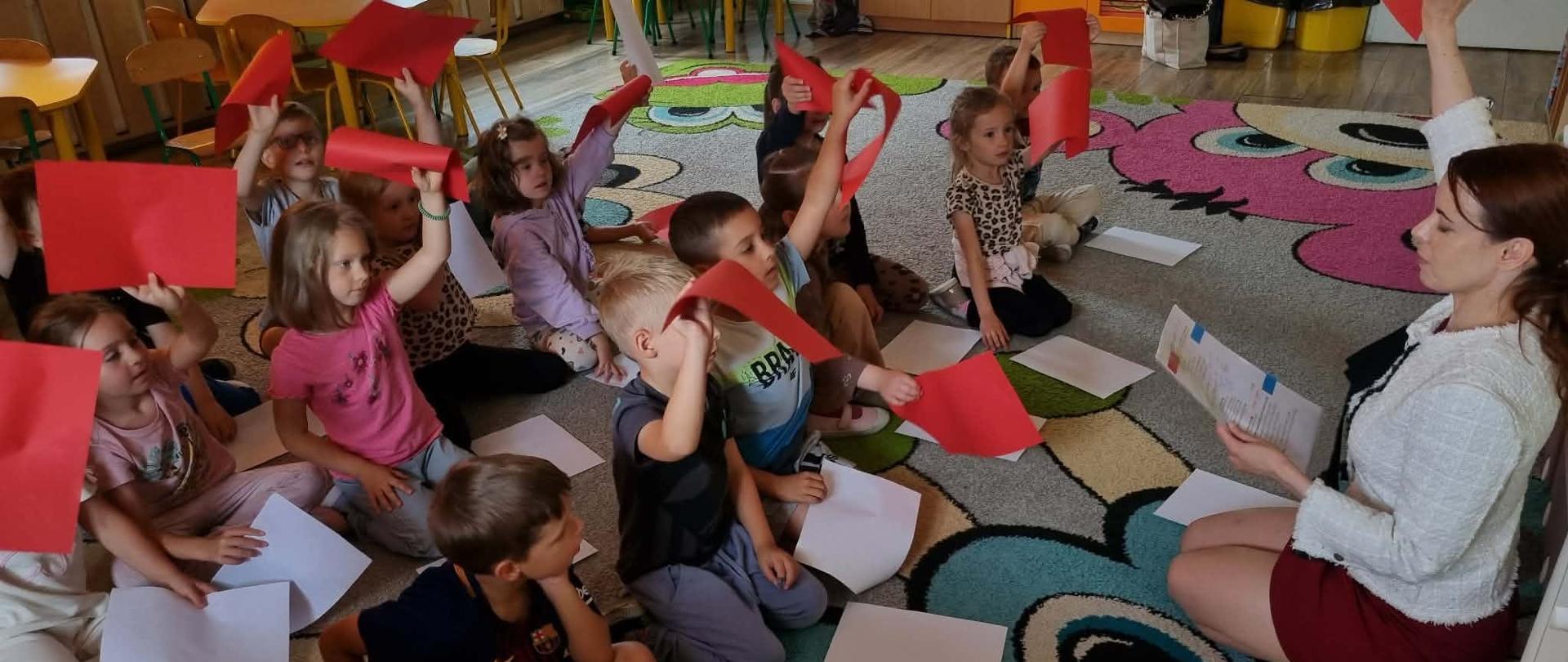 Dzieci podczas spotkania z ekspertem IPN siedzą w kole i układają flagę z białych i czerwonych kartek.