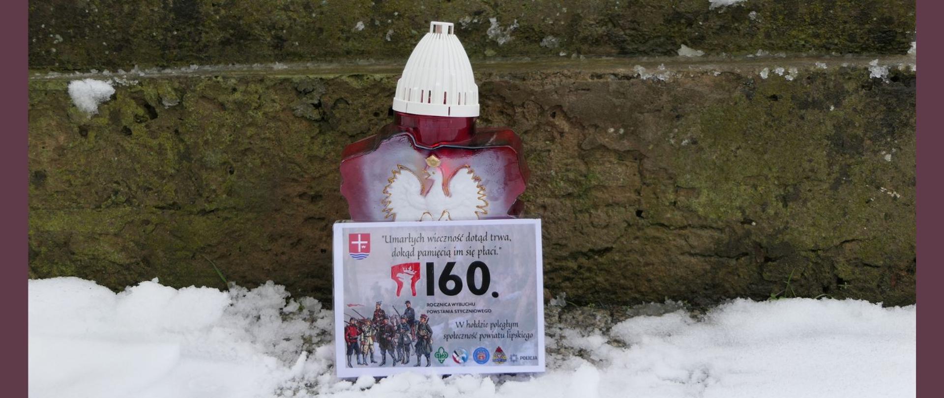 Znicz z Godłem Polski wraz z kartką intencyjną. W tle fragment mogiły powstańczej. Znicz stoi na śniegu.