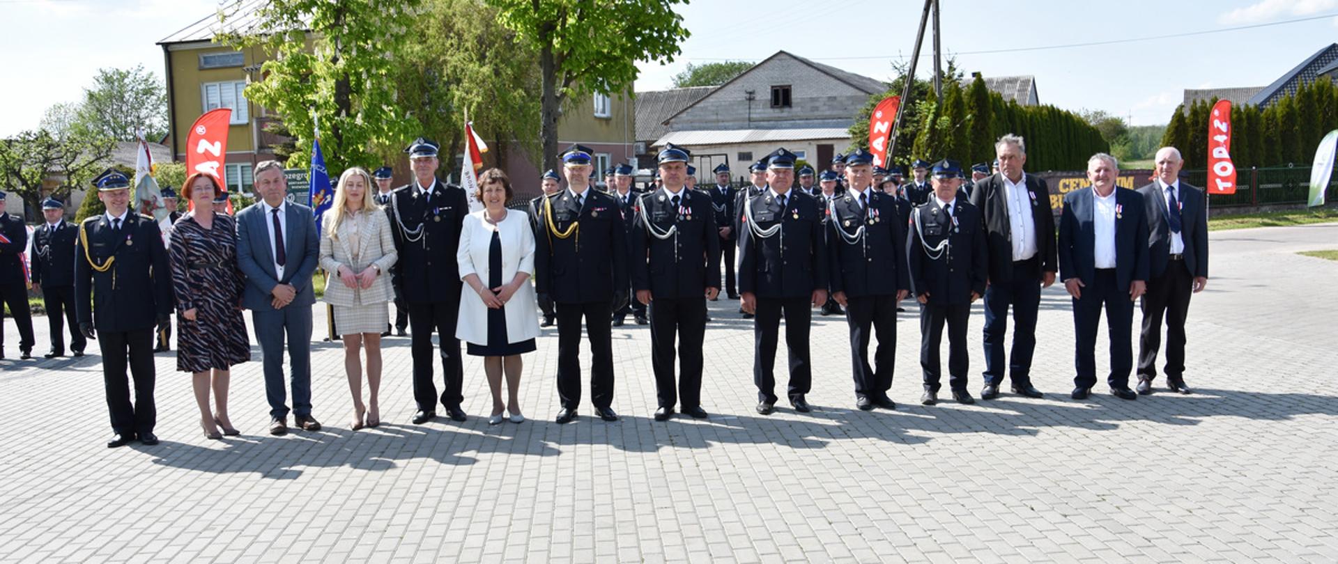 Zdjęcie grupowe na placu z betonowej kostki brukowej. Na fotografii dowództwo jednostki OSP Błonie Duże, zaproszeni goście, osoby odznaczone medalami. W tle poczty sztandarowe.