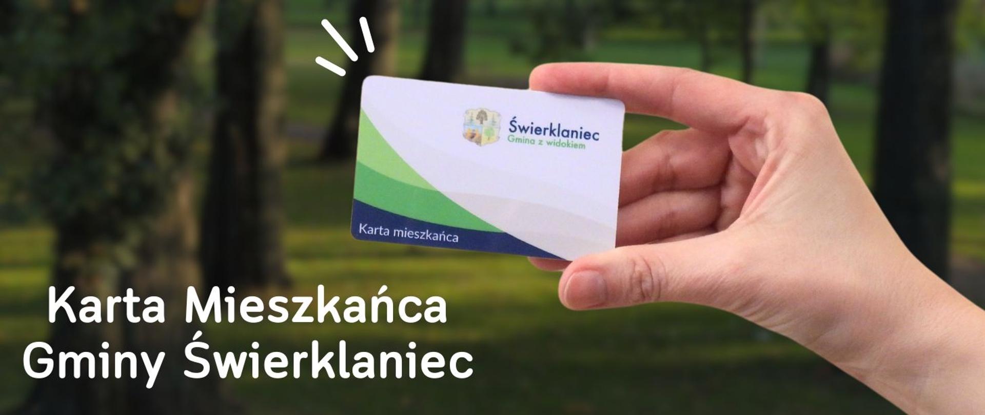 Kobieca dłoń trzyma kartę wielkości karty bankomatowej, to na tle parku z drzewami i zieloną trawą