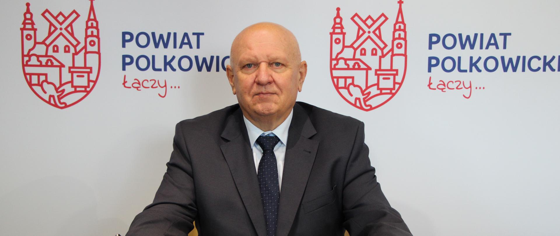 Mężczyzna siedzi przy stole w ręku ma długopis, na stole leży notes