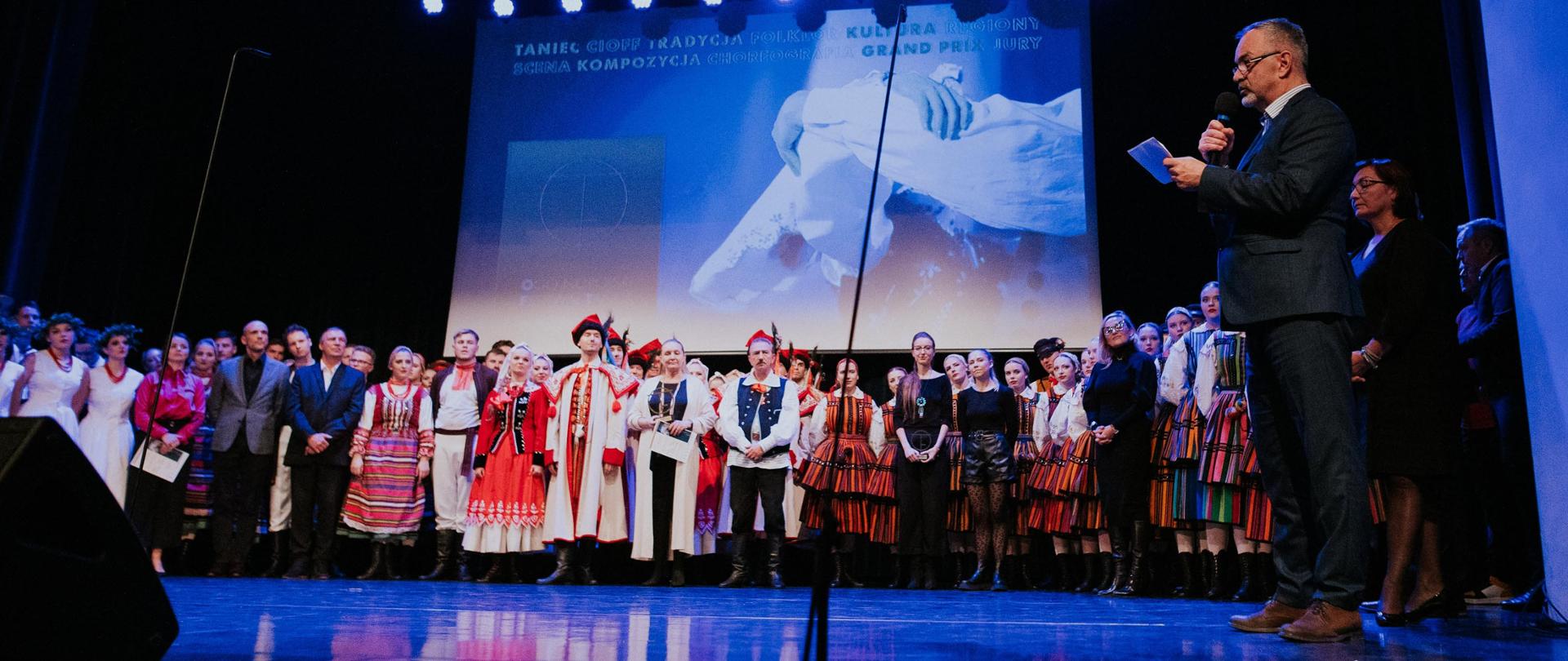 Scena sokołowskiego Ośrodka kultury, a na niej tancerze w różnych strojach ludowych, którzy uczestniczyli w koncercie finałowym. Nad nimi ekran promocyjny z grafiką festiwalową. 