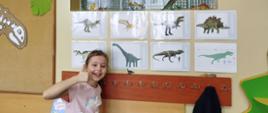 Dziewczynka stoi z uniesionym kciukiem prawej ręki przy obrazkach dinozaurów wiszących na ścianie