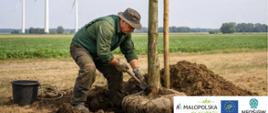 Rolnik sadzący młode drzewo na polu uprawnym; w tle farmy wiatrowe 
