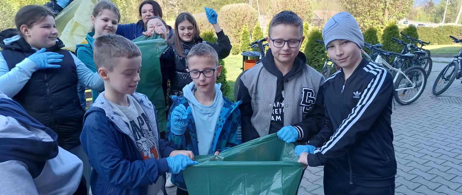 Czterej chłopcy z klasy czwartej stoją przed szkołą, trzymają wspólnie zielony worek, na rękach mają niebieskie rękawice gumowe, z lewej strony stoją cztery uczennice również trzymając zielony worek, obok stoi nauczycielka