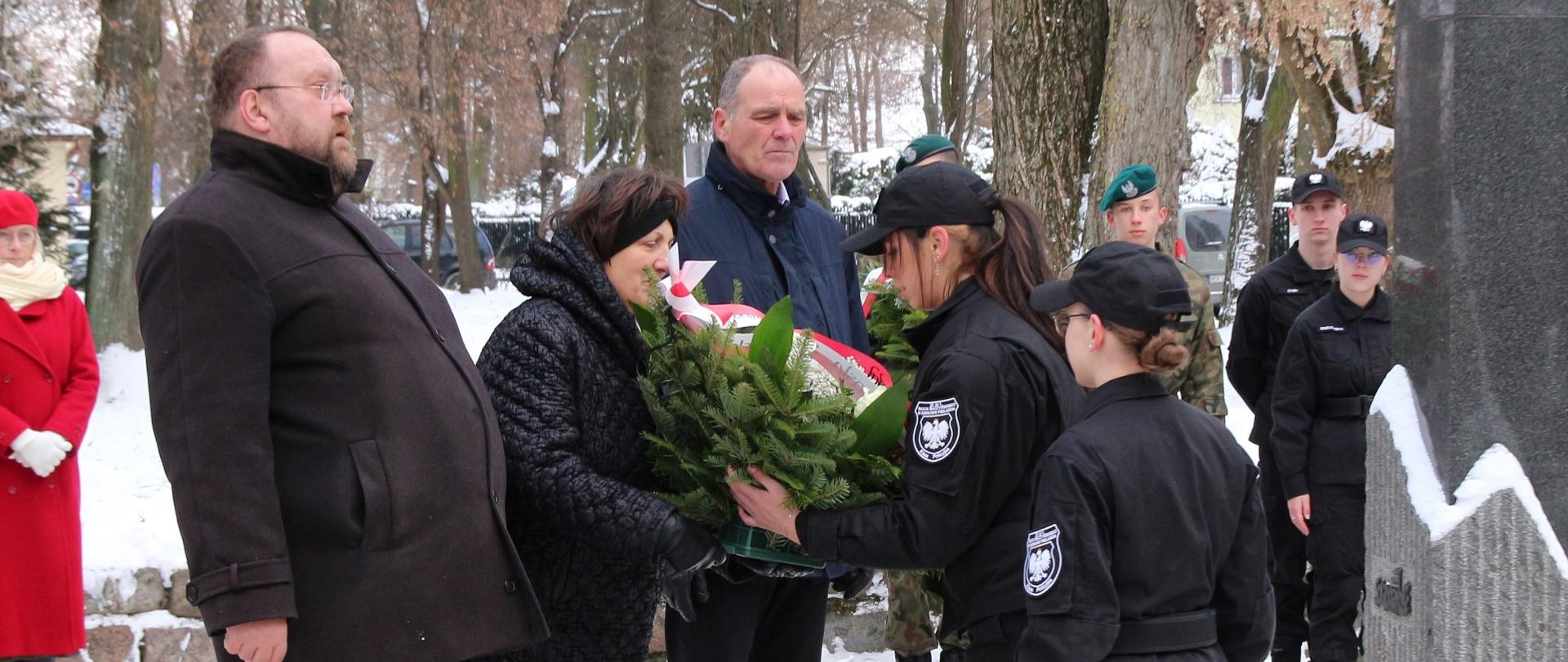 Moment Złożenia kwiatów pod pomnikiem przez Władze Powiatu Sokołowskiego. Starosta Elżbieta Sadowska przekazuje wieniec uczennicom w mundurach, które za chwilę ułożą go pod pomnikiem.