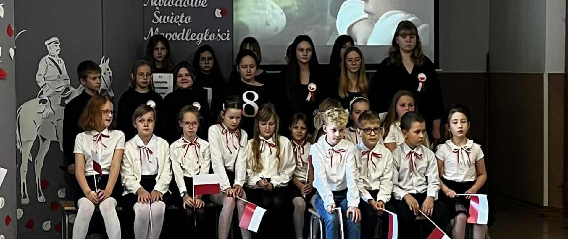Na scenie znajduje się grupa uczniów ubranych w stroje galowe. Część z nich, znajdująca się w pierwszym i drugim rzędzie - siedzi na ławkach. Uczniowie znajdujący się z tyłu - stoją. Z tyłu, za wszystkimi widnieje napis: 11 LISTOPADA. NARODOWE ŚWIĘTO NIEPODLEGŁOŚCI. 
