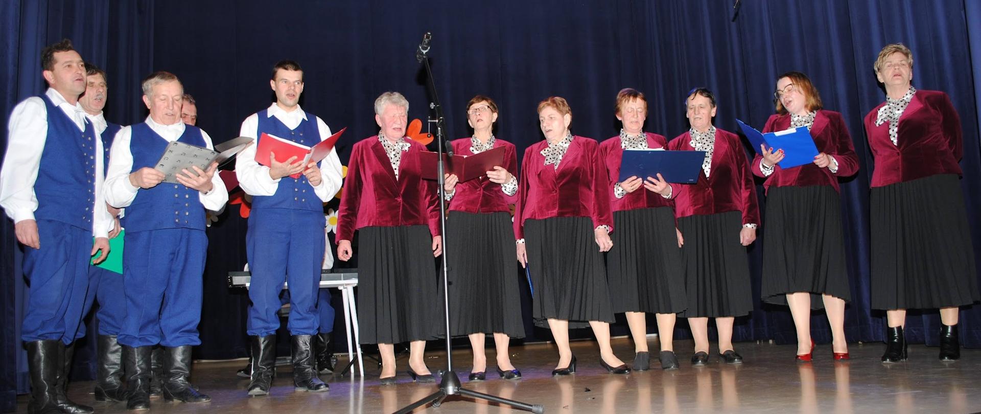siedem kobiet ubranych w różowe żakiety i czarne spódnice i kilku mężczyzn w niebieskich strojach występują na scenie 