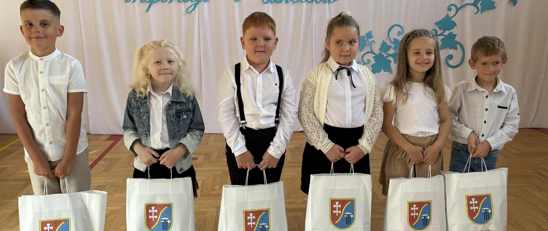 Sześcioro dzieci w białych bluzkach stoi na sali gimnastycznej. W rękach trzymają białe papierowe torby z logo Gminy Bochnia.