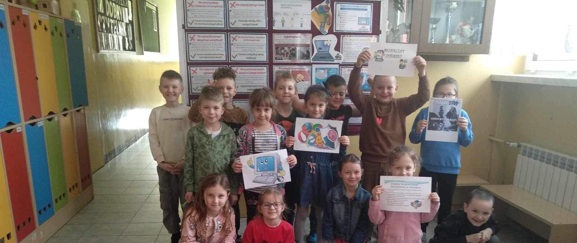 Dzieci z klasy pierwszej stoją na korytarzu, kilkoro przykuca lub leży, trzymają w rękach plakaty związane z bezpieczeństwem w sieci internetowej, za nimi gazetka informująca o akcji