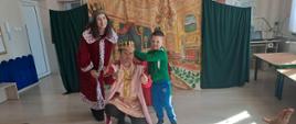 Para aktorów w królewskich strojach oraz chłopiec w zielonej bluzie, który dumnie kładzie rękę na koronie siedzącego króla. Artyści uśmiechają się do publiczności na tle bogato zdobionej dekoracji pałacowej, tworząc radosne zakończenie szkolnego przedstawienia.