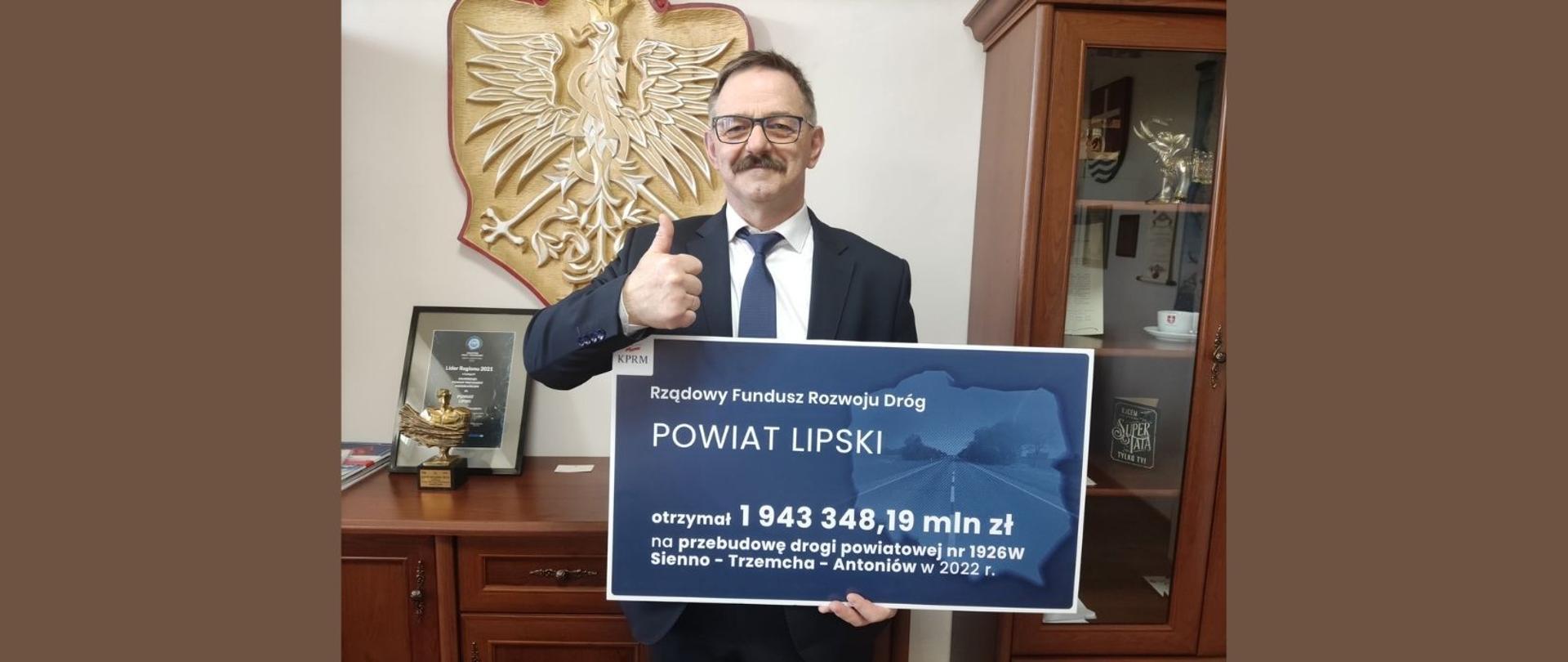 Starosta Lipski trzyma symboliczny czek, na którym widnieje kwota dofinansowania. W tle meble gabinetu Starosty oraz Godło Polski.