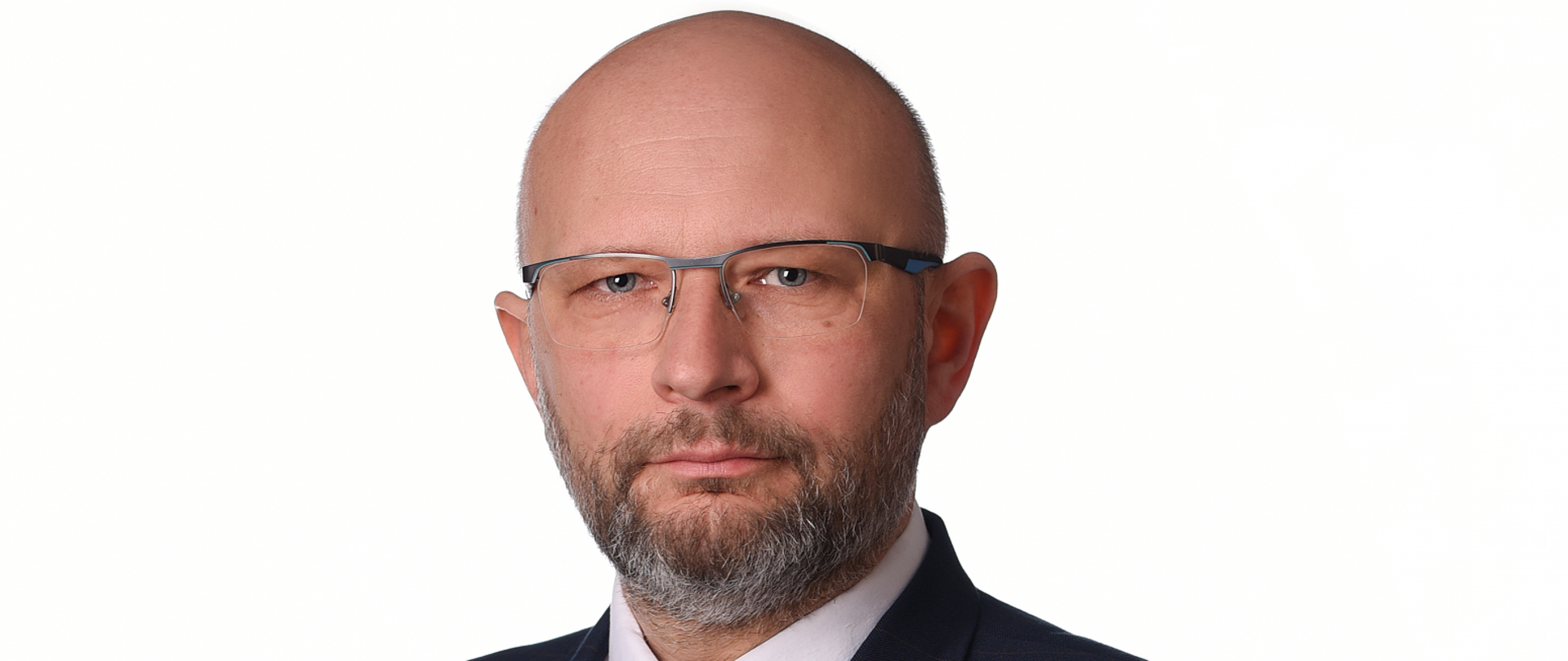 Starszy mężczyzna łysy, w okularach, lekko odstające uszy, broda i wąsy lekko szpakowate, oczy jasne. Ubrany w granatowy garnitur w delikatną brązowa kratkę, granatowy krawat.