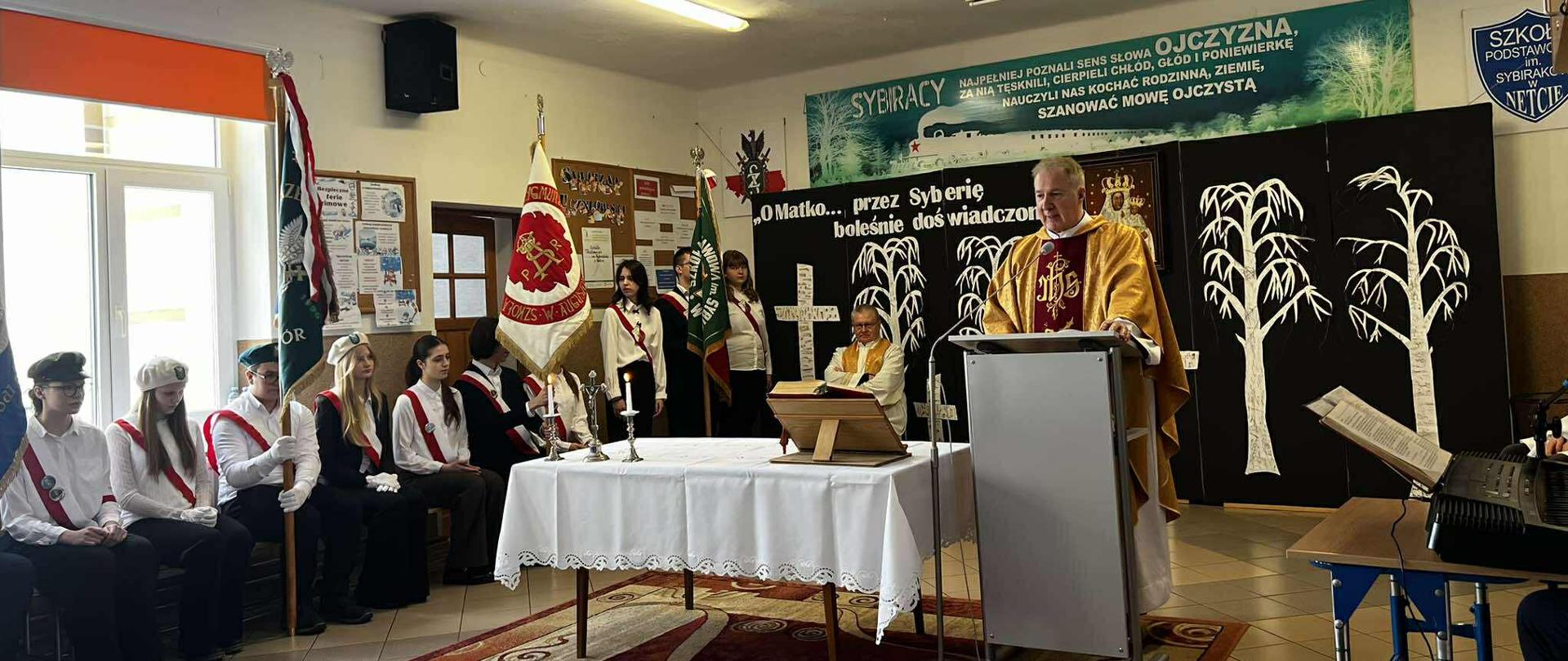 Na głównym korytarzu szkolnym odbywa się Msza Święta. Na środku stoi ołtarz oraz mównica przy której stoi ksiądz. Obok ołtarza siedzą uczniowie reprezentujący sztandary szkolne.