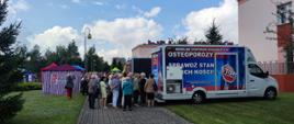 Festyn Profilaktyczno Zdrowotny w ogrodach przasnyskiego szpitala. Zdjęcie przedstawia uczestników Festynu oczekujących na badania gęstości kości w Osteobusie. W tle po prawej namiot Caritasu.