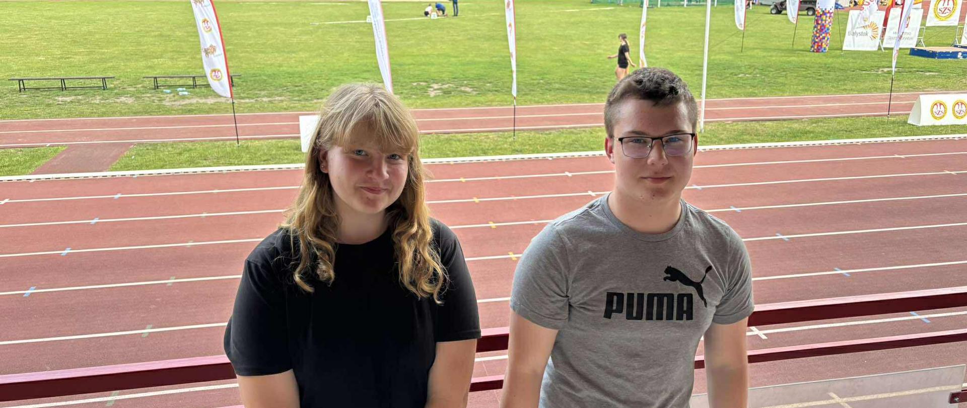 Dwoje uczniów (chłopak i dziewczyna) pozują do wspólnego zdjęcia. Oboje ubrani są w stroje sportowe. Za nimi znajduje się bieżnia stadionu lekkoatletycznego.