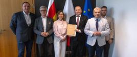 Uczestnicy spotkania pozują do pamiątkowego zdjęcia na tle flag: Podlasia, Polski i Unii Europejskiej. Mężczyzna w środku trzyma w rękach podpisaną teczkę z dokumentami, prezentując efekt zawartego porozumienia.