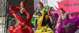 Cztery kobiety wykonują taniec na scenie w kolorowych, długich sukniach. Za nimi widać fragment baneru 100-lecie Niepodległej.