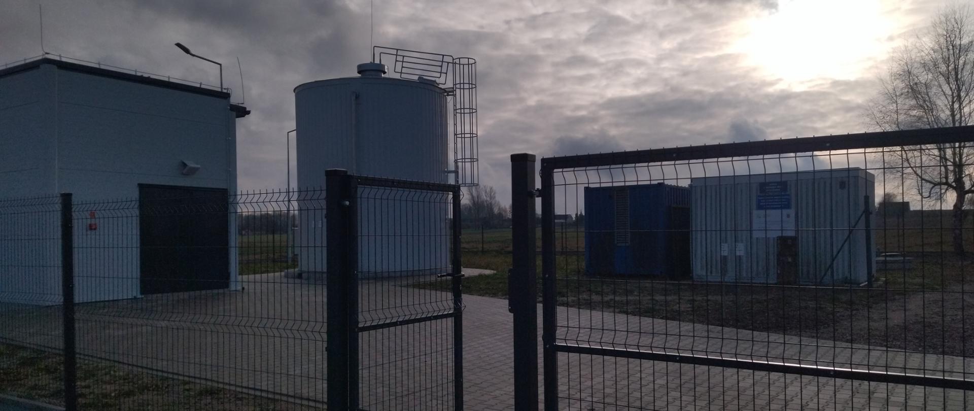Zdjęcie przedstawia industrialny obiekt otoczony metalowym ogrodzeniem z bramą. Wewnątrz ogrodzonego terenu znajdują się trzy główne struktury: duży cylindryczny zbiornik z przymocowaną drabiną, prostokątny budynek z drzwiami i lampą, oraz kontenerowa konstrukcja z niebieskim segmentem i widocznym oznaczeniem. Niebo jest zachmurzone, a przebijające się przez chmury słońce tworzy efekt dramatycznego oświetlenia.