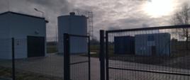 Zdjęcie przedstawia ogrodzony teren przemysłowy z dużym cylindrycznym zbiornikiem oraz mniejszą, niebiesko-białą konstrukcją, prawdopodobnie będącą budynkiem użytkowym lub magazynowym.