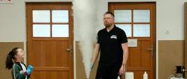 Mężczyzna w okularach i czarnej koszulce „Naukowy Patrol” stoi za stołem laboratoryjnym, z którego unosi się wysoki słup gęstego, białego dymu. Po lewej stronie stoi uśmiechnięta dziewczynka w okularach ochronnych i niebieskich rękawiczkach jednorazowych, która z zachwytem obserwuje przebieg eksperymentu. 