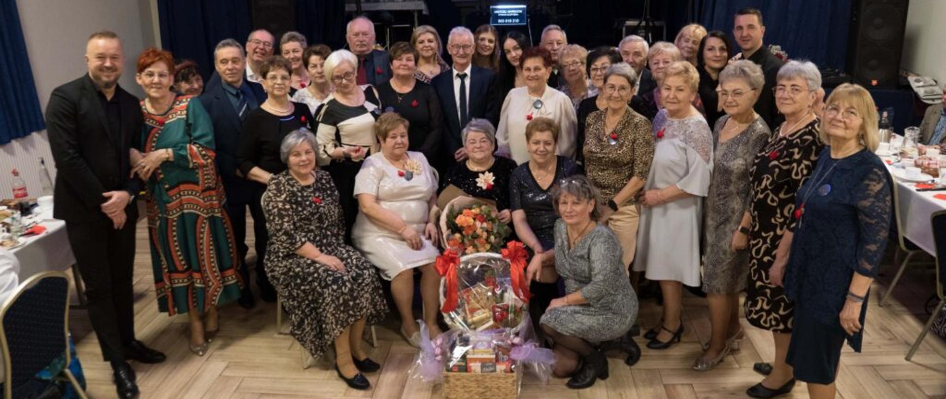 Grupa dorosłych osób pozuje do zdjęcia przed nimi kosze kwiatów i prezentów