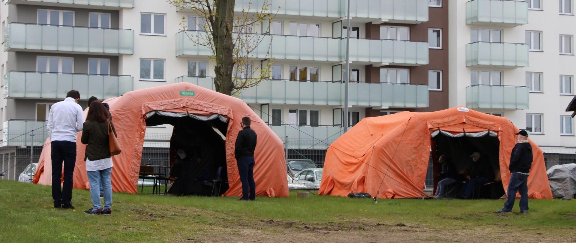 na zdjęciu znajdują się dwa pomarańczowe namioty, a w nich są ludzie, w tle widać wielomieszkaniowy blok