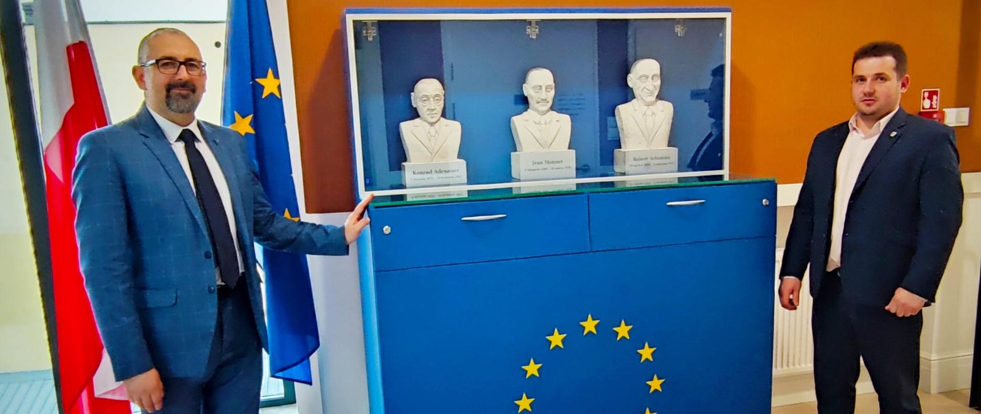 Zdjęcie przedstawia od lewej: Starostę Przasnyskiego Krzysztofa Bieńkowskiego (stojący na tle flag Rzeczypospolitej Polskiej i Unii Europejskiej) oraz Etatowego Członka Zarządu Powiatu Przasnyskiego Pawła Mostowego prezentujących gablotę poświęconą kluczowym dla powstania Unii Europejskiej postaciom. Gablota przeszklona koloru niebieskiego, u podstawy symbol Unii Europejskiej złożony z dwunastu pięcioramiennych złotych gwiazd. U góry za szkłem trzy popiersia wykonane przez artystę Andrzeja Staszewskiego z Krajewa Wielkiego. Kolejno od lewej: Konrad Adenauer, Jean Monnet, Robert Schuman.