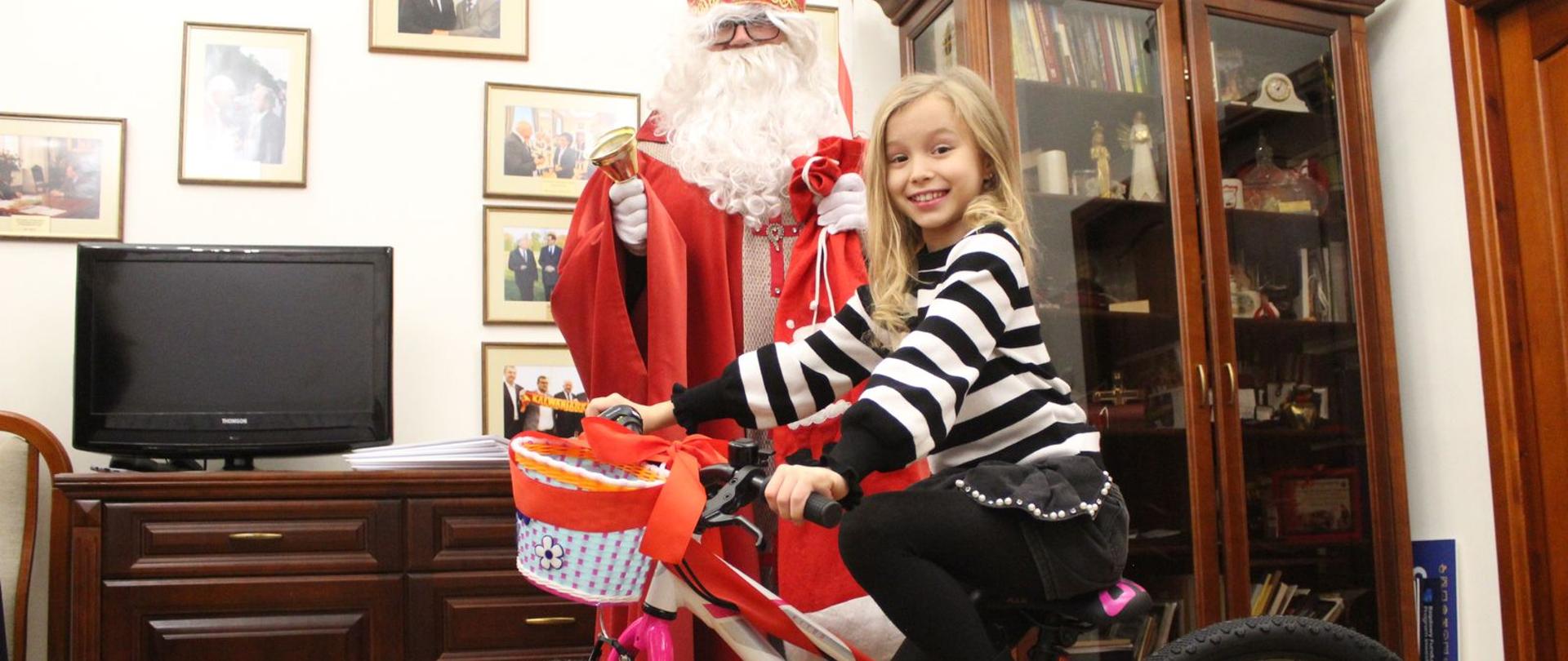 Dziewczynka na rowerze, obok święty Mikołaj. 