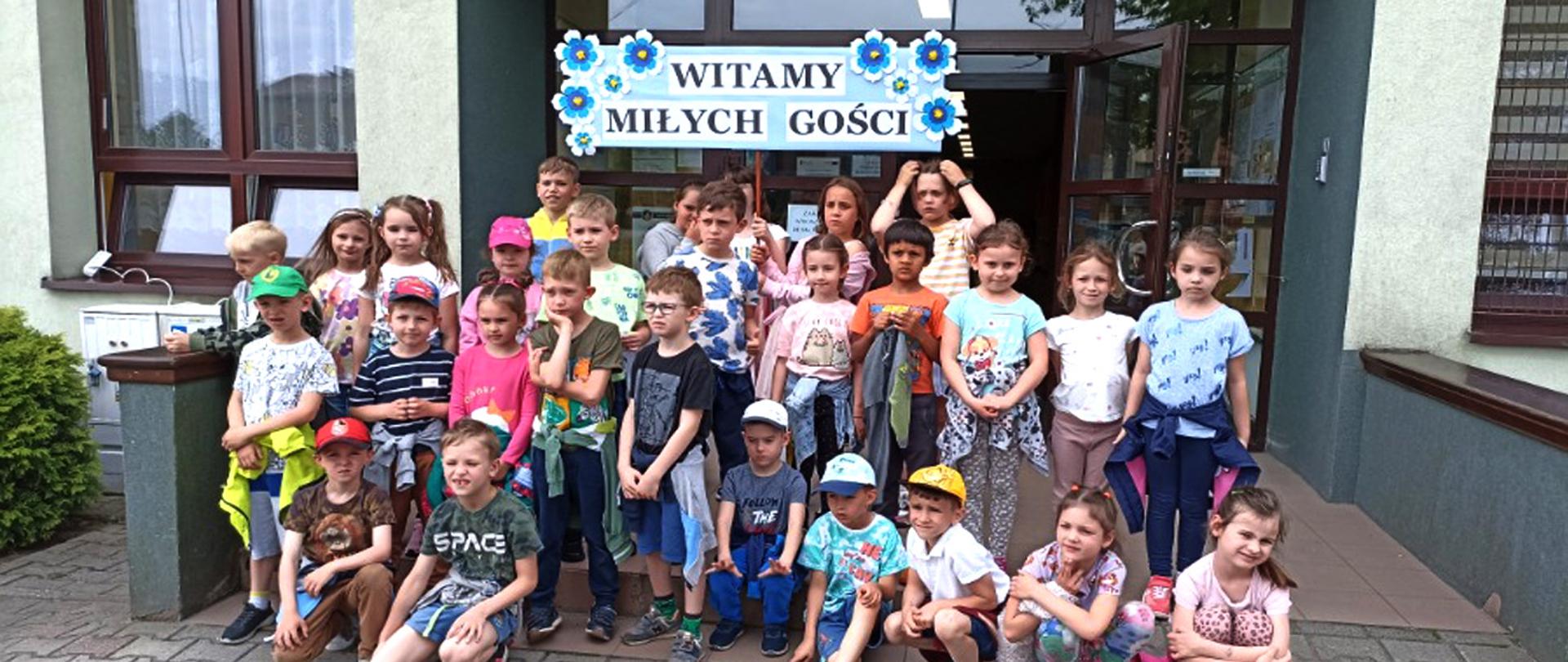 Grupa młodych uczniów trzymająca transparent witający przedszkolaków. W tle budynek szkoły.