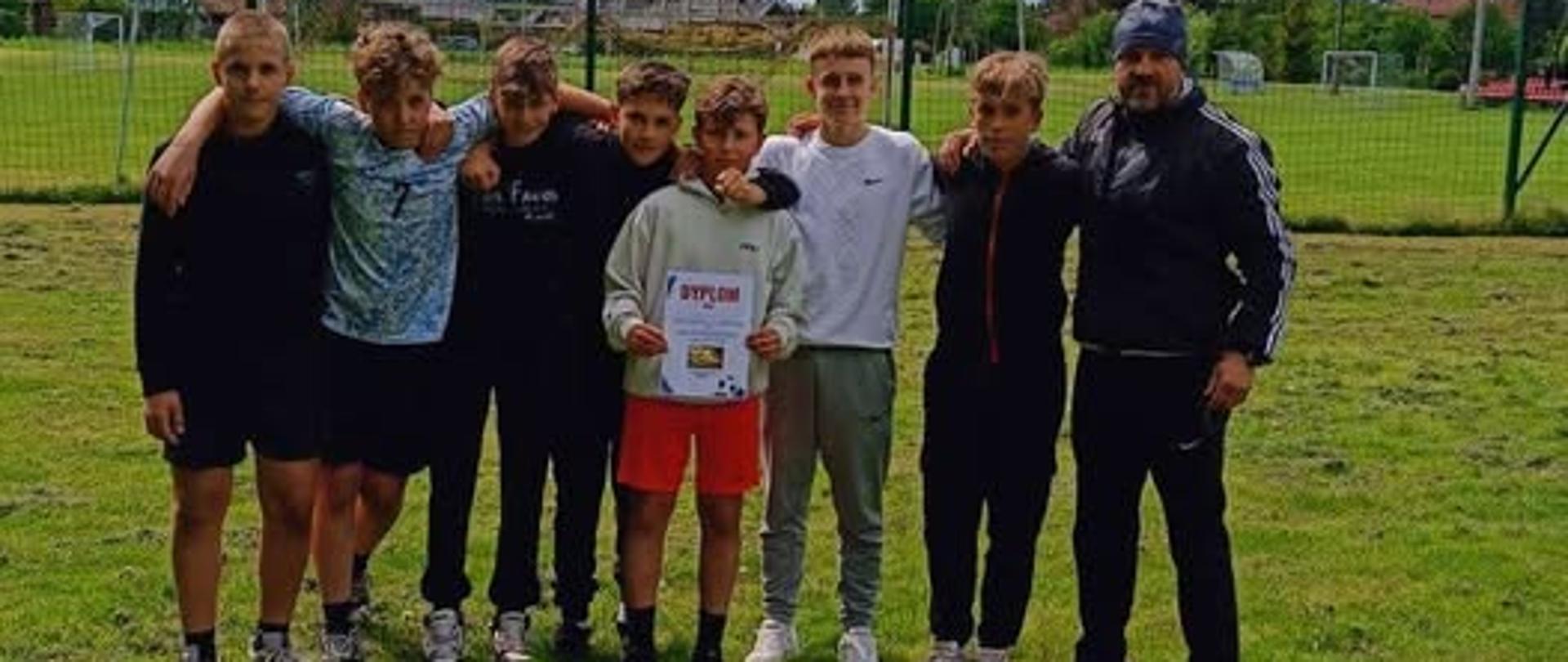Na zdjęciu widzimy ośmioosobową grupę chłopców oraz dorosłego mężczyznę stojących razem na boisku trawiastym, na tle siatki ochronnej i zielonego terenu sportowego. Chłopcy są ubrani w sportowe stroje — niektórzy w krótkich spodenkach i koszulkach, inni w dresach. Jeden z chłopców (na środku) trzyma dyplom, co sugeruje, że drużyna brała udział w zawodach i prawdopodobnie odniosła sukces.
Wszyscy stoją blisko siebie, z ramionami opartymi na barkach kolegów, tworząc zwartą, zgraną grupę. Po prawej stronie kadru stoi dorosły mężczyzna w ciemnym dresie i czapce z daszkiem – zapewne trener lub opiekun zespołu.
W tle widać fragment miejscowości, boiska, bramki piłkarskie i latarnie. Niebo jest częściowo zachmurzone, ale widać też przebłyski błękitu, co nadaje zdjęciu pogodną atmosferę. Całość emanuje sportowym duchem i dumą z osiągnięcia.