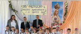 Pamiątkowe zdjęcie dużej grupy maluchów w identycznych strojach (chłopcy w szelkach i muchach, dziewczynki w kolorowych spódniczkach tiulowych). Za nimi na scenie stoją dwie opiekunki oraz dwaj mężczyźni. Jedno z dzieci trzyma dyplom dla grupy „Misie”.