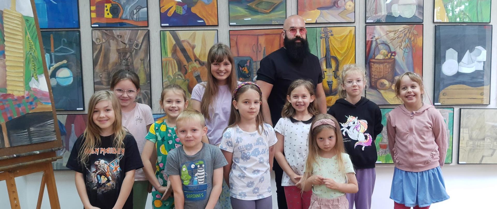 Na zdjęciu dzieci, uczestnicy zajęć artystycznych z nauczycielem panem Marcinem Bondarowiczem, wszyscy stoją na tle obrazów prezentowanych w sali Ogniska plastycznego. 