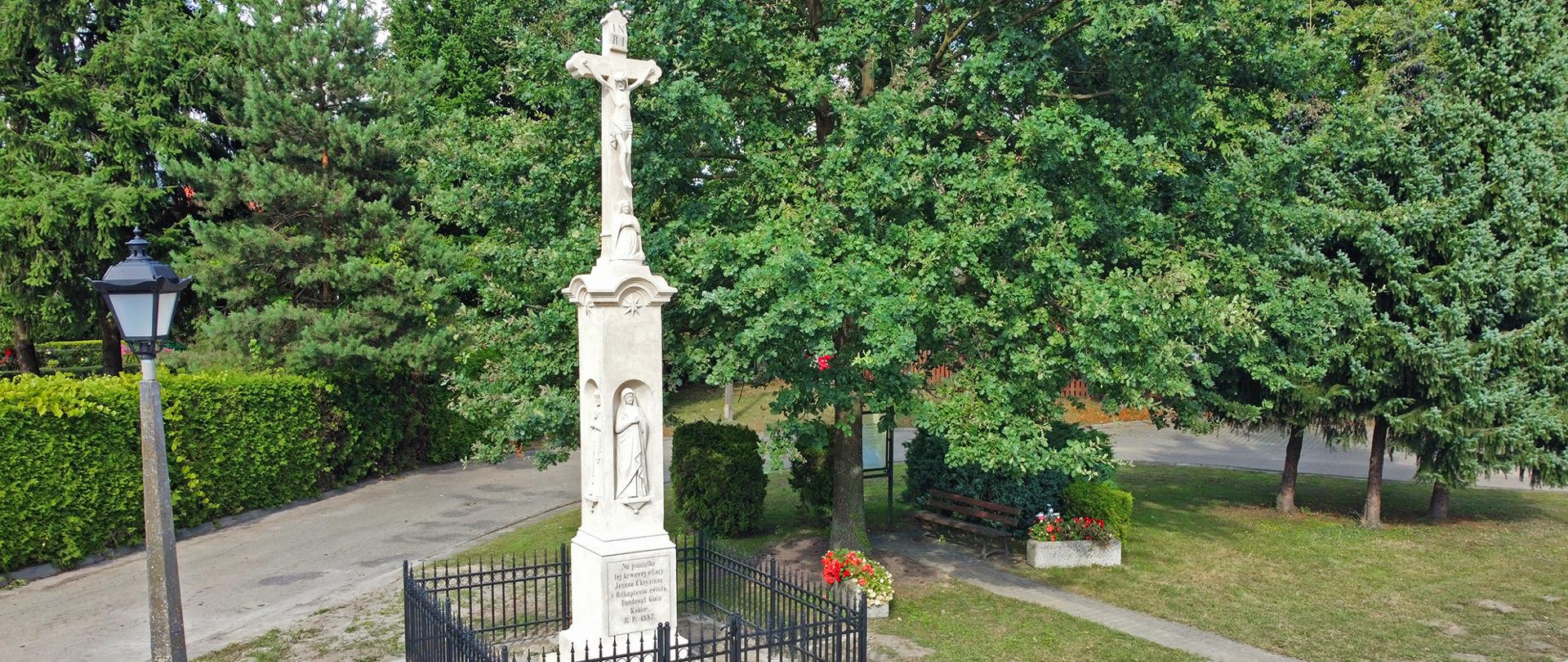Krzyż z piaskowca ogrodzony czarnym płotem. Obok latarnia i zielone drzewa.