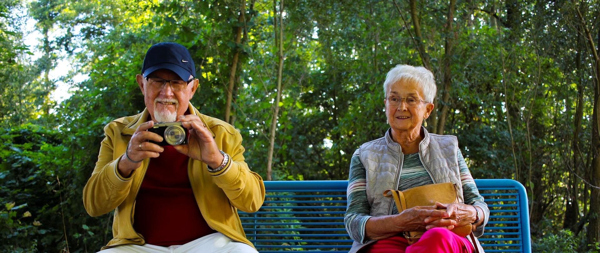 Dwie osoby siedzą na ławce w parku (zdjęcie wykonane na zewnątrz). Jedna z osób trzyma w ręce aparat fotograficzny.