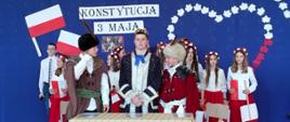 Przy stole stoją trzej uczniowie odgrywający role twórców konstytucji 3 maja, przebrani w stroje szlacheckie, w środku uczeń przebrany za króla, za nimi dziewczynki ubrane w białe koszule i czerwone długie spódnice, w biało-czerwonych wiankach na głowie