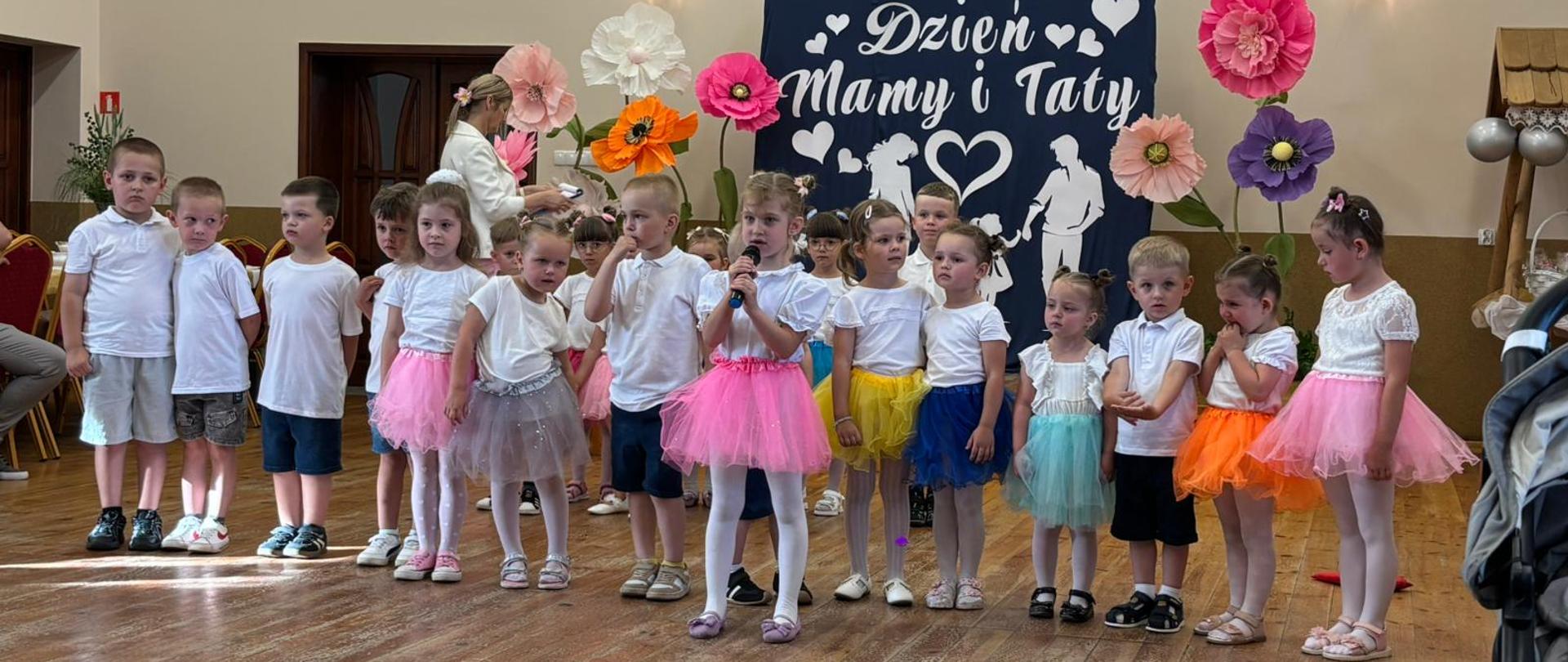 Zdjęcie przedstawia salę o jasnych ścianach i podłodze drewnianej. Na pierwszym planie widać grupę dzieci w wieku przedszkolnym. Dziewczynki w kolorowych spódniczkach i białych bluzeczkach, chłopcy w krótkich spodenkach i białych bluzeczkach. Za nimi kobieta stojąca bokiem o jasnych włosach, w kremowej bluzce. W tle ścianka a na niej napis Dzień Mamy i Taty, z boku duże kolorowe kwiaty. Jedna dziewczynka trzyma w rękach mikrofon. 